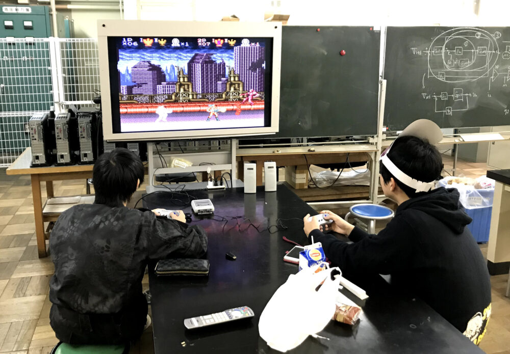 理科室のような教室で、黒いテーブルの上に、スーパーファミコンを置いて、ゲームをする学生たち